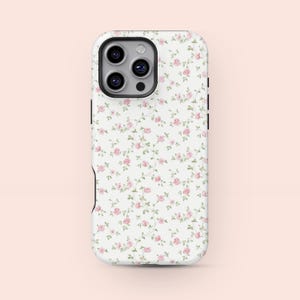Può includere: Una custodia per telefono bianca con un motivo floreale di piccole rose rosa e foglie verdi. La custodia ha un ritaglio per la fotocamera e pulsanti laterali neri.