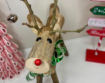 May include: A reindeer figurine crafted from natural wood branches, featuring a bright red bead nose, two black bead eyes, and a green and white striped ribbon tied around its neck. The antlers are made from thin, bare twigs.