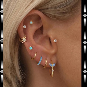 Op de afbeelding: Een oor versierd met meerdere gouden oorbellen. De collectie omvat kleine hoepels, studs met heldere en turquoise stenen en een sterrenpatroon. De sieraden creëren een gelaagde en stijlvolle look, met een verscheidenheid aan vormen en kleuren.