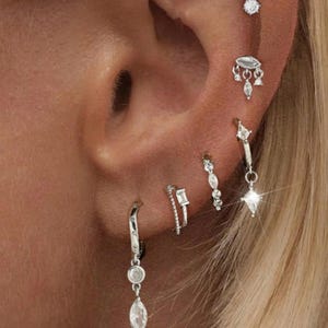 May include: Close-up of an ear with multiple silver earrings. The earrings have various designs, including dangling teardrops, geometric bars, and sparkling studs. The ear is set against a backdrop of blonde hair.