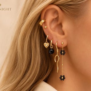Könnte beinhalten: Eine Sammlung goldfarbener Ohrringe, bestehend aus einer Ohrklemme, einem kleinen Stecker, einem Creolen-Herzanhänger, einer Creole mit schwarzer Perle, einem gewellten Ohrhänger und einer langen Kette mit zwei schwarzen Blumen. Text TTHENIGHT ist sichtbar.