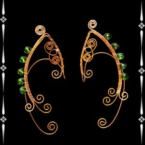 May include: A pair of handcrafted ear cuffs with a golden wire design. The ear cuffs feature green gemstone beads and decorative swirls. The background is black, highlighting the intricate details of the jewelry.