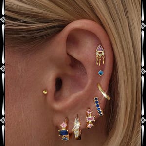 May include: Close-up of an ear adorned with multiple gold earrings. The earrings feature various designs, including a triangle with dangling chains, a small blue stud, a curved hoop, and several small hoops with colorful gems.