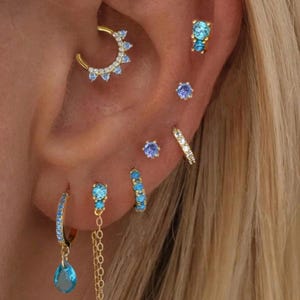 Könnte beinhalten: Nahaufnahme eines Ohrs, das mit mehreren goldenen und blauen Edelsteinohrringen geschmückt ist. Die Kollektion umfasst Creolen, Ohrstecker und einen hängenden Tropfenohrring. Der Schmuck weist verschiedene blaue Edelsteine und eine goldene Oberfläche auf.