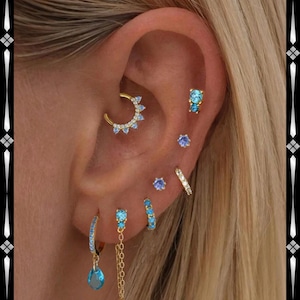 Può includere: Un orecchio è adornato da una selezione di orecchini color oro con pietre preziose blu. La collezione comprende un cerchio, borchie e un orecchino pendente con una gemma a forma di goccia. Diversi piercing mostrano la varietà di gioielli.