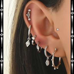 Può includere: Un orecchio adornato da diversi orecchini d'argento. Gli orecchini includono un ciondolo a forma di diamante pendente, piccoli cerchi e una barra curva con pietre trasparenti. È presente anche un piccolo orecchino a bottone.