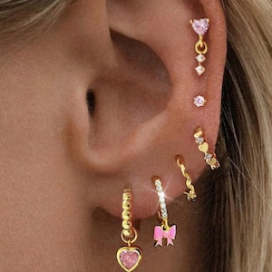 Può includere: Primo piano di un orecchio adornato da molteplici orecchini d'oro. Gli orecchini presentano vari design, tra cui ciondoli a forma di cuore con gemme rosa, un fiocco e piccoli borchie. I gioielli sono ambientati su uno sfondo di capelli biondi.