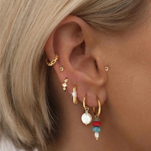 Könnte beinhalten: Ein Ohr ist mit mehreren goldenen Ohrringen geschmückt, darunter ein Kettenglied-Reifen, kleine Ohrstecker und ein Paar hängende Reifen mit Perlen und bunten Perlen. Die Ohrringe zeigen eine Vielzahl von Designs und Texturen.
