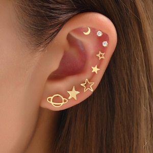 Op de afbeelding: Een verzameling goudkleurige oorbellen in verschillende hemelse vormen, waaronder een halve maan, sterren en een planeet. De oorbellen worden op een oor getoond, waarbij de verschillende ontwerpen en maten worden getoond. De oorbellen zijn een stijlvol accessoire.