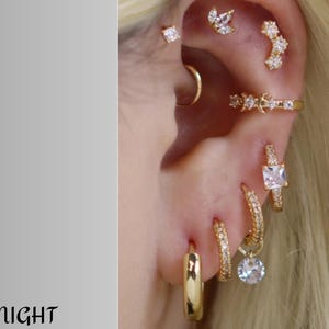 Puede incluir: Primer plano de una oreja adornada con múltiples pendientes dorados. Los pendientes incluyen aros, tachuelas y un brazalete de cartílago, todos con brillantes piedras de circonita cúbica.