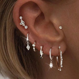 Puede incluir: Primer plano de una oreja adornada con múltiples pendientes de plata. La oreja presenta un delicado trepador de oreja, pequeños aros y pendientes colgantes con detalles brillantes. La joyería añade un toque de elegancia y brillo.
