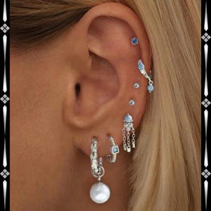 Pode incluir: Close-up de uma orelha adornada com múltiplos brincos de prata. Os brincos incluem pequenos pregos, argolas com pedras azuis quadradas e desenhos de correntes penduradas com detalhes azuis e uma grande pérola branca.