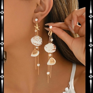 Puede incluir: Pendientes colgantes dorados con conchas marinas, perlas y un dije de estrella de mar. Los pendientes tienen un diseño delicado con múltiples hebras y pequeñas perlas redondas. Las conchas marinas son blancas con acentos marrones.