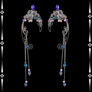 May include: A pair of silver ear cuffs with floral and scrollwork designs. Each cuff features a cluster of small flowers and gemstones in shades of blue, purple, and clear. Dangling chains with blue and purple teardrop-shaped beads complete the design.