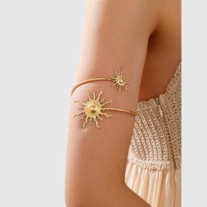 Peut inclure: Un bracelet de bras en métal doré orné de deux motifs en forme de soleil. Le design se compose d'une fine bande flexible qui s'enroule autour du haut du bras, avec un emblème solaire plus grand à une extrémité et un plus petit à l'autre.