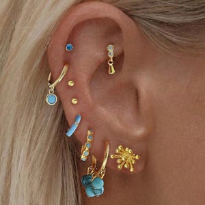 Può includere: Primo piano di un orecchio adornato con molteplici orecchini dorati. Gli orecchini includono piccoli borchie, cerchi con dettagli turchesi e design pendenti. Alcuni orecchini presentano pietre preziose blu, creando un look elegante.
