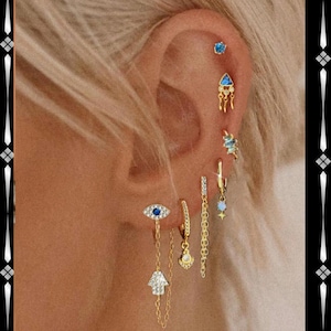 May include: Close-up of an ear with multiple gold earrings. The earrings showcase diverse designs, including a blue eye, a hand, a triangle, and a star. Some earrings have dangling chains and small gemstones. The ear is set against a backdrop of blonde hair.