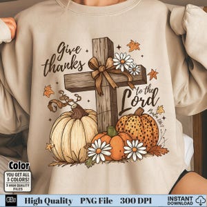 May include: Beige sweatshirt with a fall-themed graphic. The design includes a wooden cross with a brown bow, pumpkins, daisies, and the text "Give thanks to the Lord". The image also includes the text "Psalm 136:7".