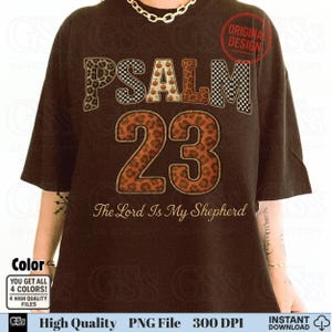 May include: Dark brown t-shirt with the text "PSALM 23" in a mix of leopard print, checkered, and pumpkin patterns. The phrase "The Lord Is My Shepherd" is below the numbers. The shirt has a round "Original Design" stamp.