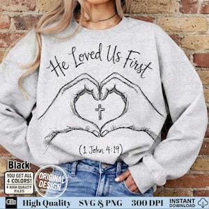 May include: A light gray sweatshirt with the text "He Loved Us First" in a script font. Below the text is a drawing of two hands forming a heart shape with a cross in the center. The text "(1 John 4:19)" is below the heart.