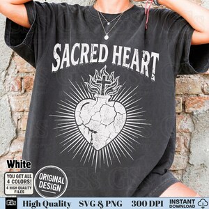 Op de afbeelding: Een zwart t-shirt met de witte tekst "SACRED HEART" boven een witte afbeelding van een hart met een kruis en vlammen. Het ontwerp heeft een verweerde, vintage uitstraling. Extra tekst bevat "White", "ORIGINAL DESIGN" en "High Quality SVG & PNG".