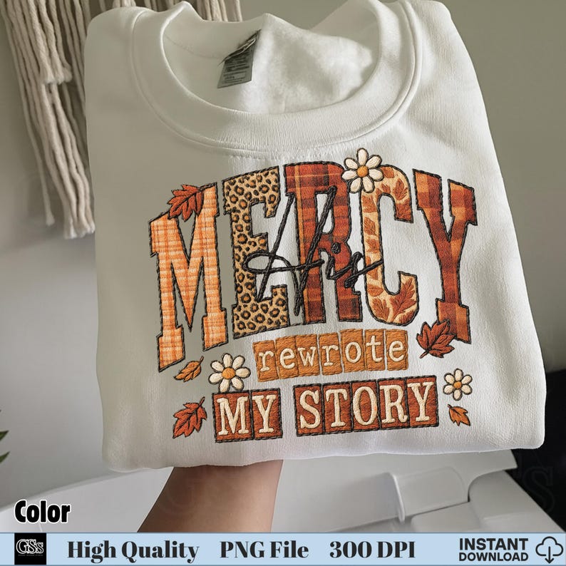 White folded sweatshirt with fall colors “Mercy His Rewrote My Story” Christian shirt design
