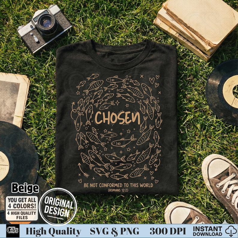Flat lay of a black t-shirt on grass with beige "Chosen" design, vintage camera, and books.