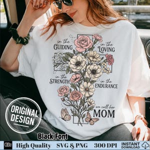 以下が含まれることがあります： 白いTシャツに花の十字架のデザイン。デザインにはピンクと白の花、蝶、そして「in the Guiding, Loving, Strength, Endurance, we call her MOM」という文字が含まれています。シャツには「Original Design」と「Black Font」の文字があります。