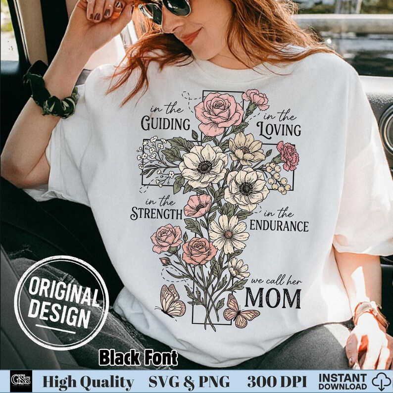 以下が含まれることがあります： 白いTシャツに花の十字架のデザイン。デザインにはピンクと白の花、蝶、そして「in the Guiding, Loving, Strength, Endurance, we call her MOM」という文字が含まれています。シャツには「Original Design」と「Black Font」の文字があります。