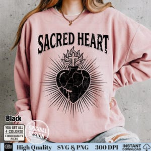 Op de afbeelding: Een lichtroze sweatshirt met de tekst "SACRED HEART" in een versleten lettertype. Daaronder een zwarte afbeelding van een hart met een kruis en vlammen. De afbeelding bevat ook tekst: "Black", "YOU GET ALL 4 COLORS!" en "ORIGINAL DESIGN".