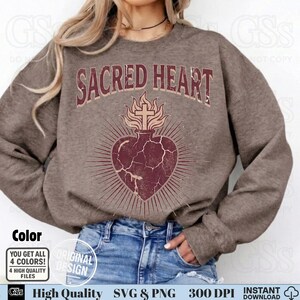 Op de afbeelding: Een bruine sweatshirt met de tekst "SACRED HEART" boven een hartontwerp met een kruis en vlammen. Het hart is dieprood met stralende lijnen. De sweatshirt heeft een neutrale kleur, geschikt voor diverse stijlen.