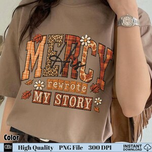 Hand holding shirt with autumn leaf and flower themed embroidery “Mercy His Rewrote My Story”