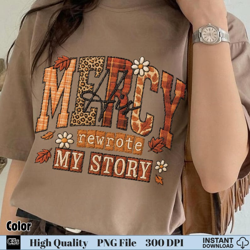 Hand holding shirt with autumn leaf and flower themed embroidery “Mercy His Rewrote My Story”