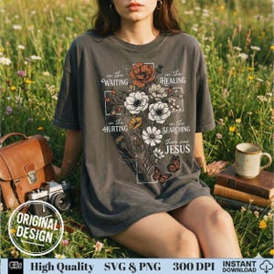 May include: Dark gray t-shirt featuring a floral cross design with the words "in the waiting, healing, hurting, searching, there was Jesus." The design includes flowers and butterflies. The shirt has a relaxed fit and short sleeves.