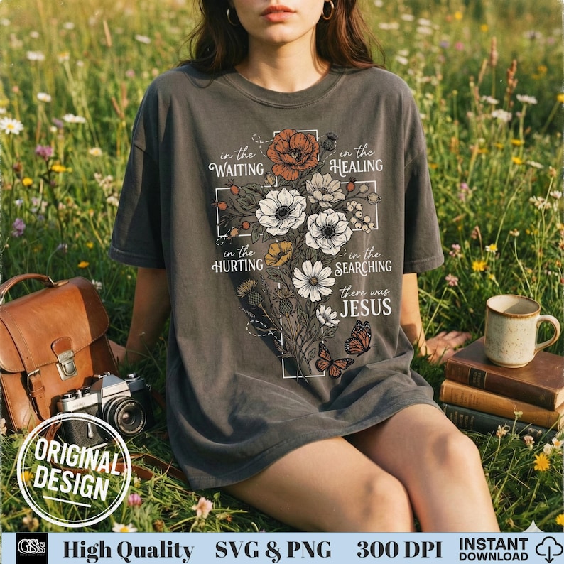 May include: Dark gray t-shirt featuring a floral cross design with the words "in the waiting, healing, hurting, searching, there was Jesus." The design includes flowers and butterflies. The shirt has a relaxed fit and short sleeves.