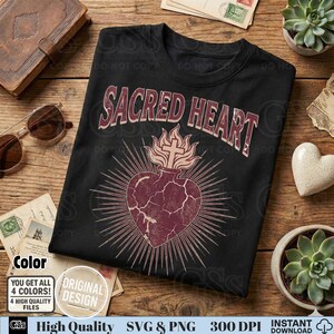 Op de afbeelding: Zwart T-shirt met de tekst "SACRED HEART" in een vintage stijl. Het ontwerp toont een hart met een kruis en vlammen, omringd door stralen. Het T-shirt heeft een klassieke ronde hals.