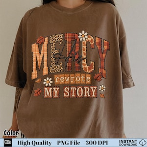 Puede incluir: Camiseta marrón con las palabras "MERCY" y "rewrote MY STORY" en un diseño decorativo con temática otoñal. Las letras están rellenas de estampado de leopardo, cuadros y motivos florales. La camiseta tiene mangas cortas.