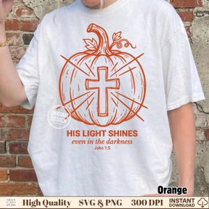 Könnte beinhalten: Ein weißes T-Shirt mit einer orangefarbenen Grafik eines Kürbisses mit einem Kreuz in der Mitte. Der Text lautet: HIS LIGHT SHINES even in the darkness John 1:5. Das Design enthält strahlenförmige Linien und einen runden Stempel ORIGINAL DESIGN.