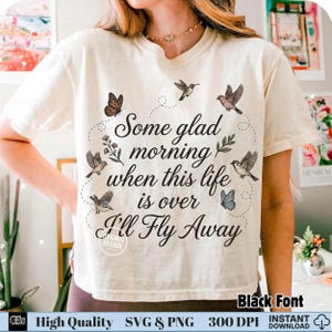 以下が含まれることがあります： 「Some glad morning when this life is over I'll Fly Away」の黒い筆記体で書かれたクリーム色のTシャツ。デザインには蝶と鳥が含まれています。シャツはクルーネックと半袖です。