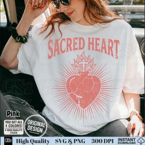 Op de afbeelding: Wit T-shirt met een roze grafisch ontwerp. Het ontwerp toont de woorden "SACRED HEART" boven een hart met een kruis en stralende lijnen, met een verweerde, vintage look. Extra tekst bevat "Pink", "Original Design" en "High Quality SVG & PNG".