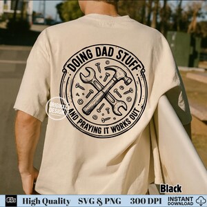 May include: A beige short-sleeved t-shirt featuring a black circular graphic on the back. The design includes a crossed hammer and wrench surrounded by various screws and nuts, with the text DOING DAD STUFF AND PRAYING IT WORKS OUT.