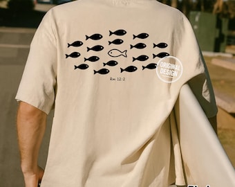 May include: A beige short-sleeve t-shirt featuring a black graphic on the back. The design displays a school of small fish swimming in one direction, with a single outlined fish swimming in the opposite direction. The text Rm 12:2 is printed below.