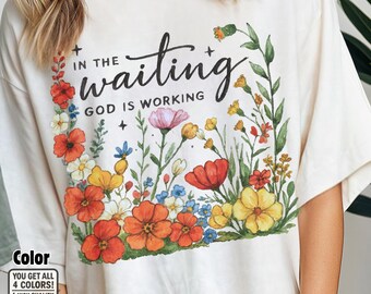 Puede incluir: Camiseta color crema con un diseño floral y la frase "In the waiting, God is working". El arreglo floral incluye flores rojas, naranjas, amarillas, rosas y azules con follaje verde. La camiseta tiene un corte holgado.