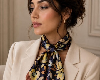 May include: A square neck scarf featuring a dark navy blue background with a floral pattern of yellow and light blue flowers and green leaves. The scarf has a solid brown border and is tied in a knot around the neck over a cream-colored blazer.