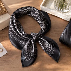 May include: A black and white silk scarf with a leopard print design. The scarf is knotted and lies on a wooden surface, next to a small black purse and a framed picture. A gold ring and a card with text are also visible.