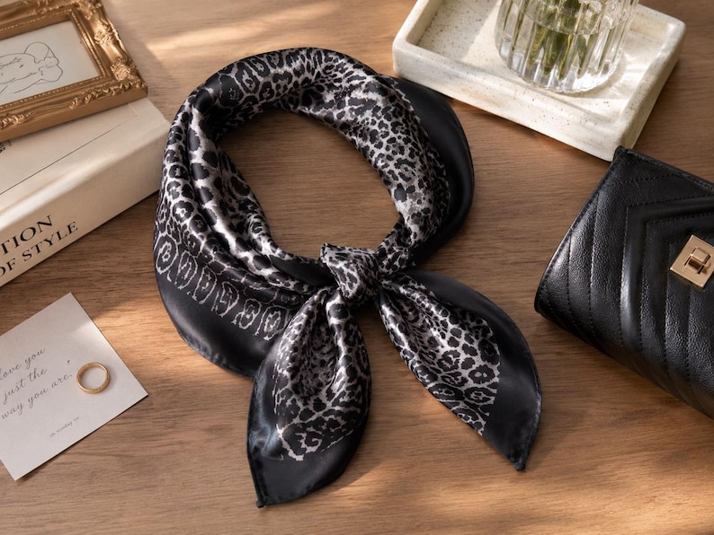 May include: A black and white silk scarf with a leopard print design. The scarf is knotted and lies on a wooden surface, next to a small black purse and a framed picture. A gold ring and a card with text are also visible.