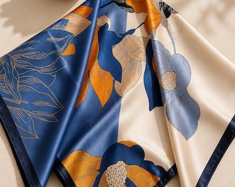 May include: A square silk scarf with a floral design. The scarf features a color block design with navy blue, cream, and orange hues. The floral pattern includes large stylized flowers and leaves. The scarf is folded, showing the different sections and colors.
