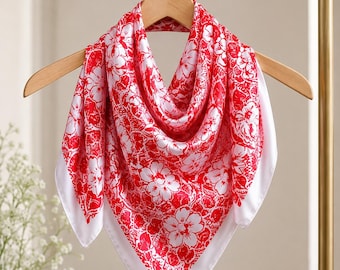 May include: A square red and white floral patterned scarf hanging on a wooden hanger. The scarf has a white border and a repeating floral design in red and white. The scarf is made of a silky material.