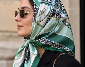 May include: A silk scarf in shades of green, white, and black, patterned with geometric and equestrian motifs, is tied around the head. The scarf is paired with a black blazer and gold-rimmed sunglasses. A gold chain strap is visible.