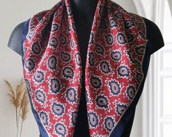 May include: A red paisley patterned scarf with a navy blue border is draped over a black mannequin. The scarf features a repeating floral design in white and navy blue. The scarf is made of a silky material.
